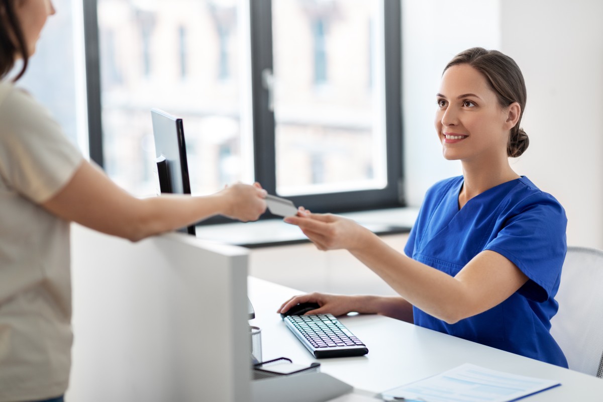 A healthcare professional in blue scrubs receives a card from a patient across a reception desk in a clinic.