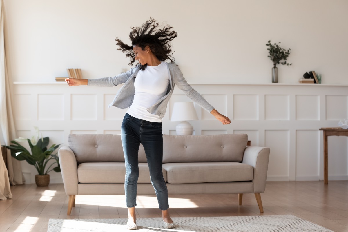 Woman joyfully dancing alone in a bright living room with white walls and a gray couch.