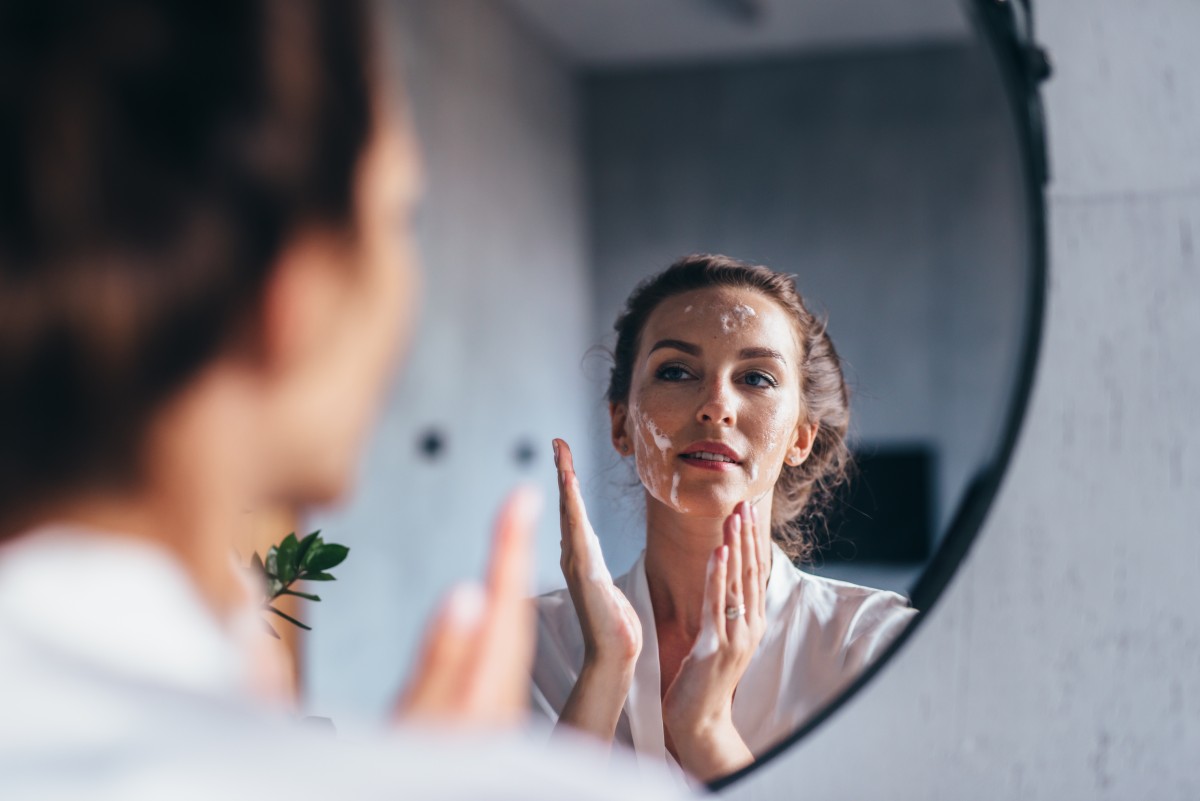 A woman examines her reflection in a round mirror, applying facial cleanser or cream to her face.