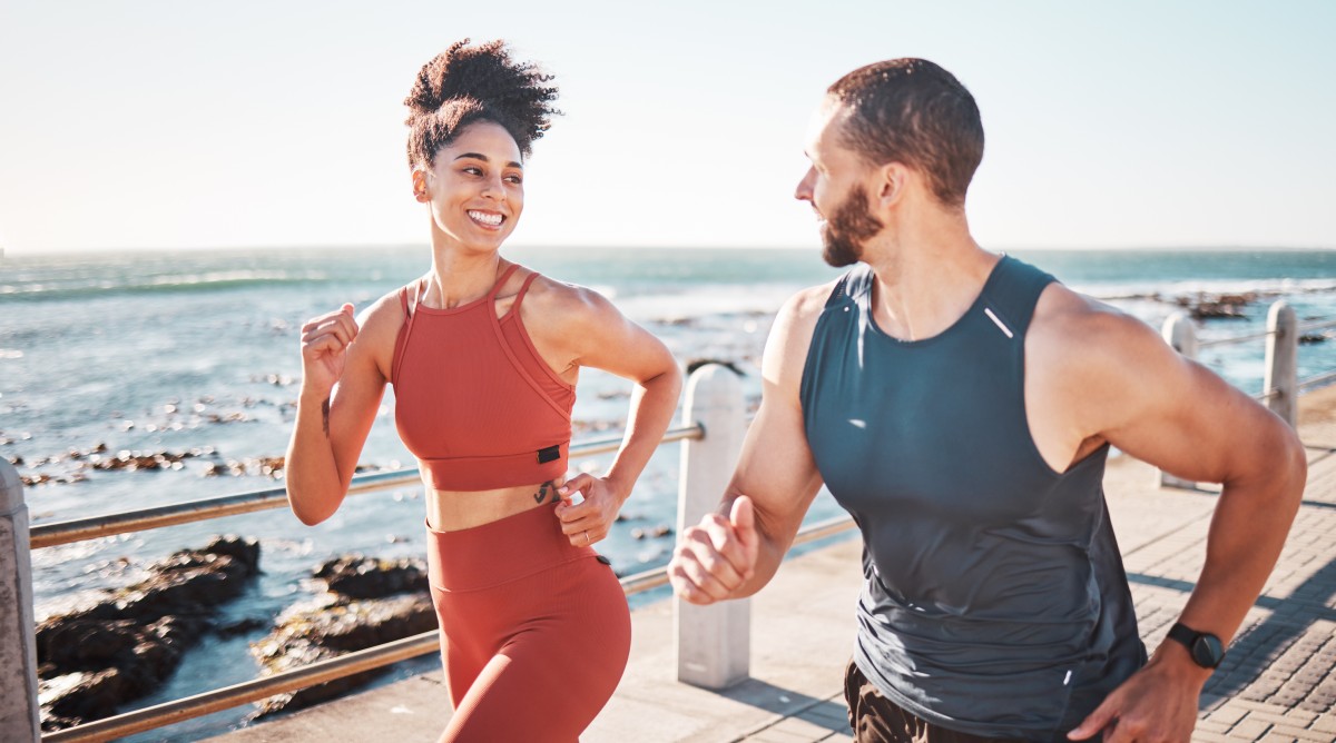 Two people jogging together on a seaside promenade, smiling and talking, with the ocean in the background.