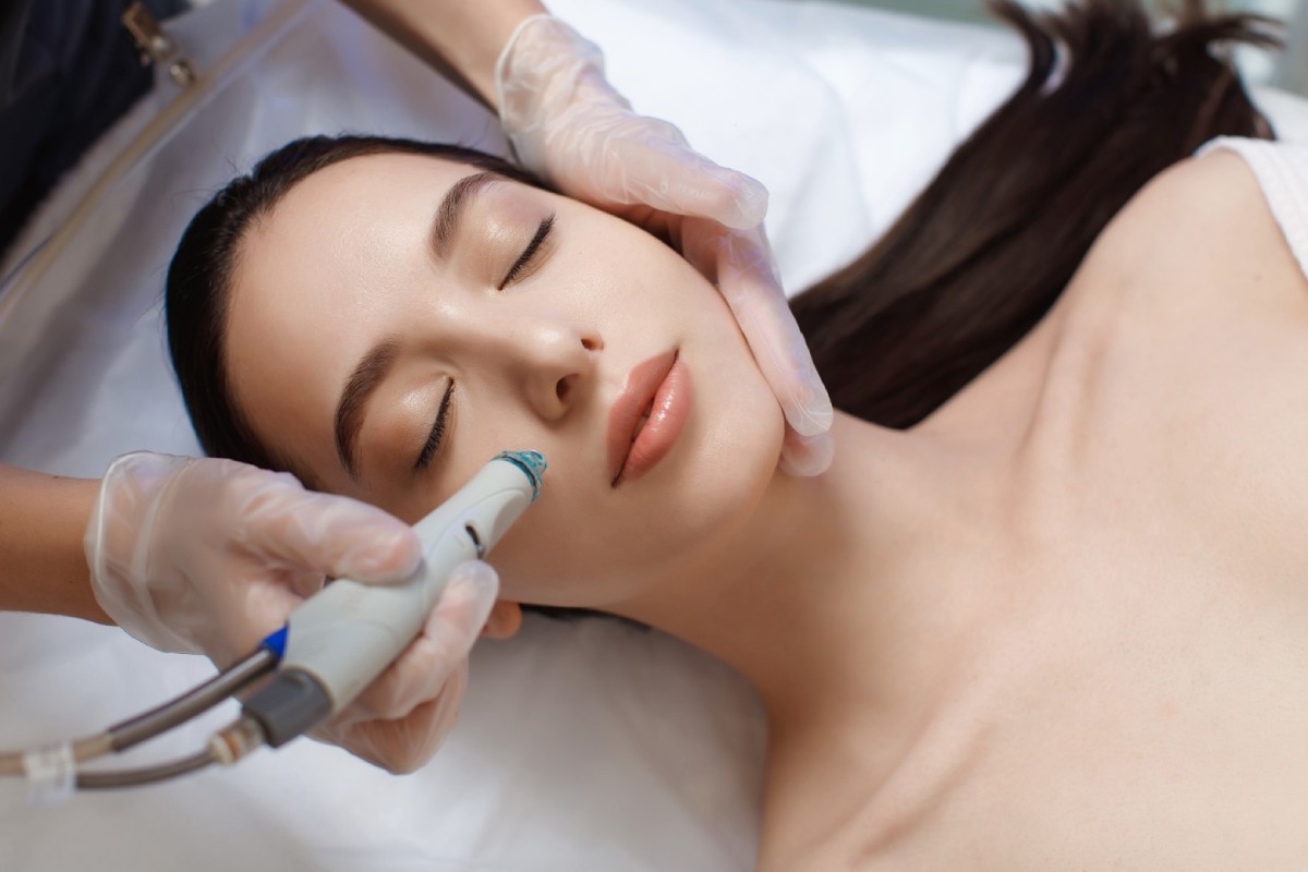 A woman receiving a facial treatment with a specialized device, lying down while a professional in gloves applies the treatment.