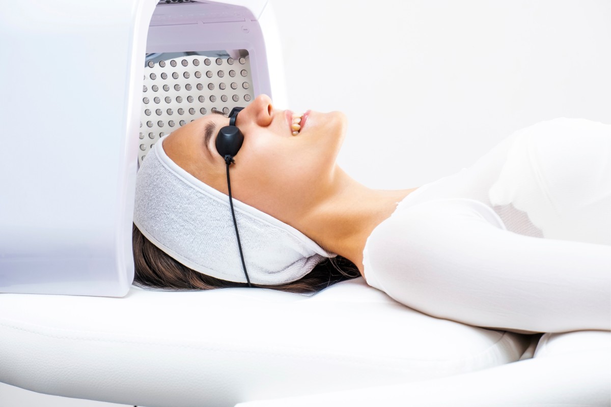 A woman lying under a led light therapy machine wearing protective goggles and a headband in a spa setting.