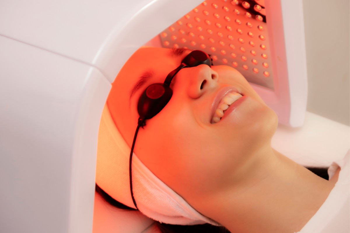 A woman wearing protective goggles smiles while receiving a red light facial therapy treatment.