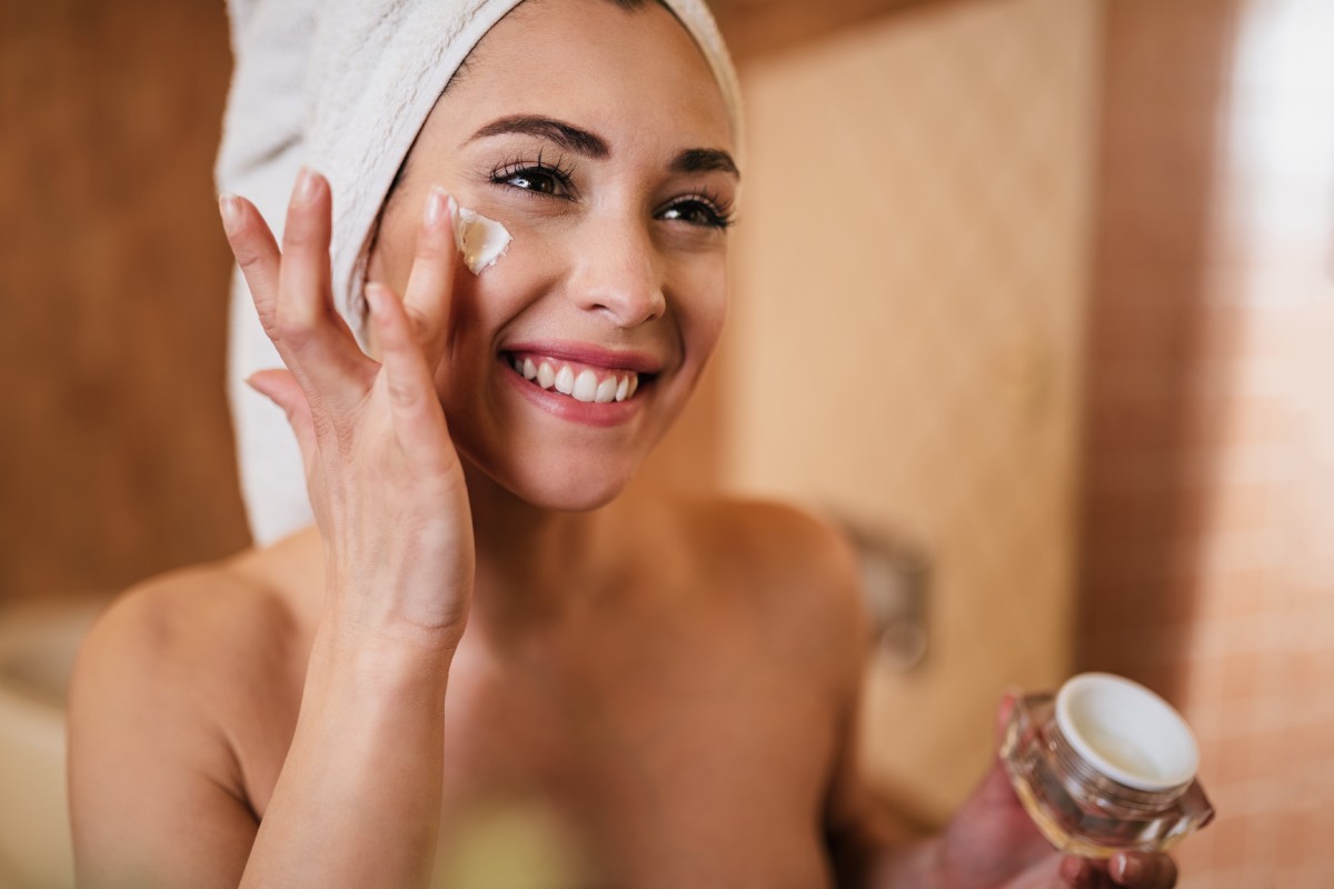 A smiling woman with a towel on her head applies facial cream in a warmly lit bathroom.