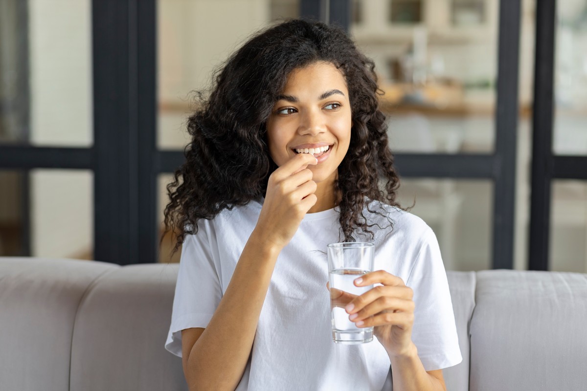 A young woman with curly hair, smiling, holding a glass of water and taking supplements while sitting on a couch in a well-lit room.
