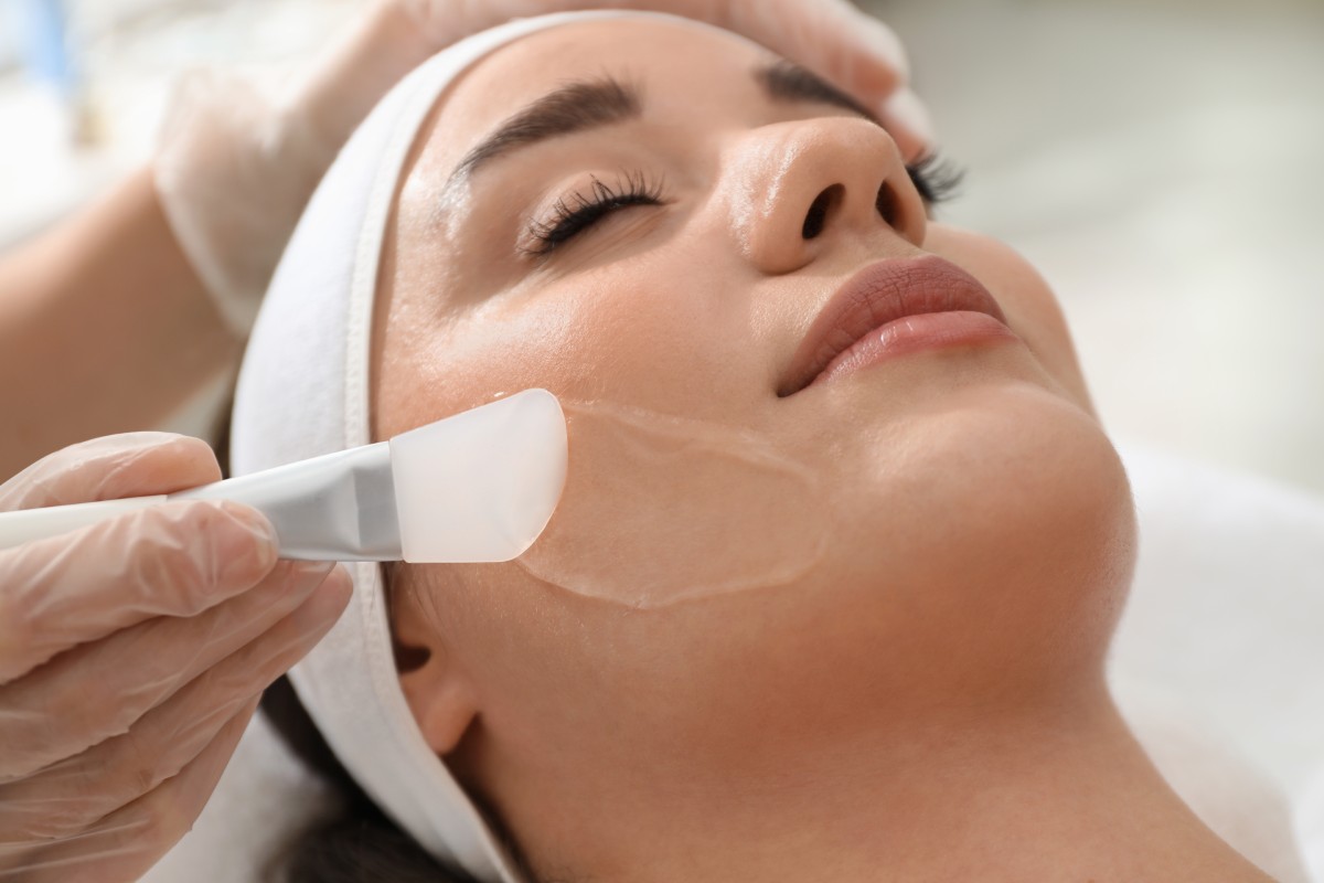 Aesthetician applying a facial mask on a woman's face using a brush, in a spa setting.