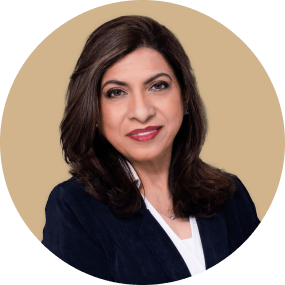 Professional headshot of Dr Dinar Sajan - a smiling woman with shoulder-length brown hair, wearing a navy blazer and white top, against a beige background.