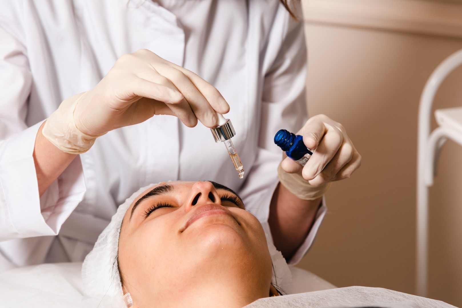 Aesthetician applying serum using a dropper on a female client's face during a skincare treatment in a clinic.