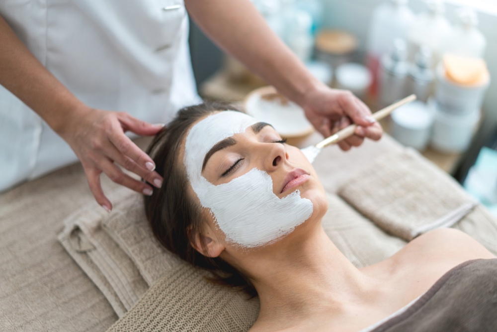 A woman lies with eyes closed, receiving a facial treatment with a brush application of a white mask in a serene spa setting.