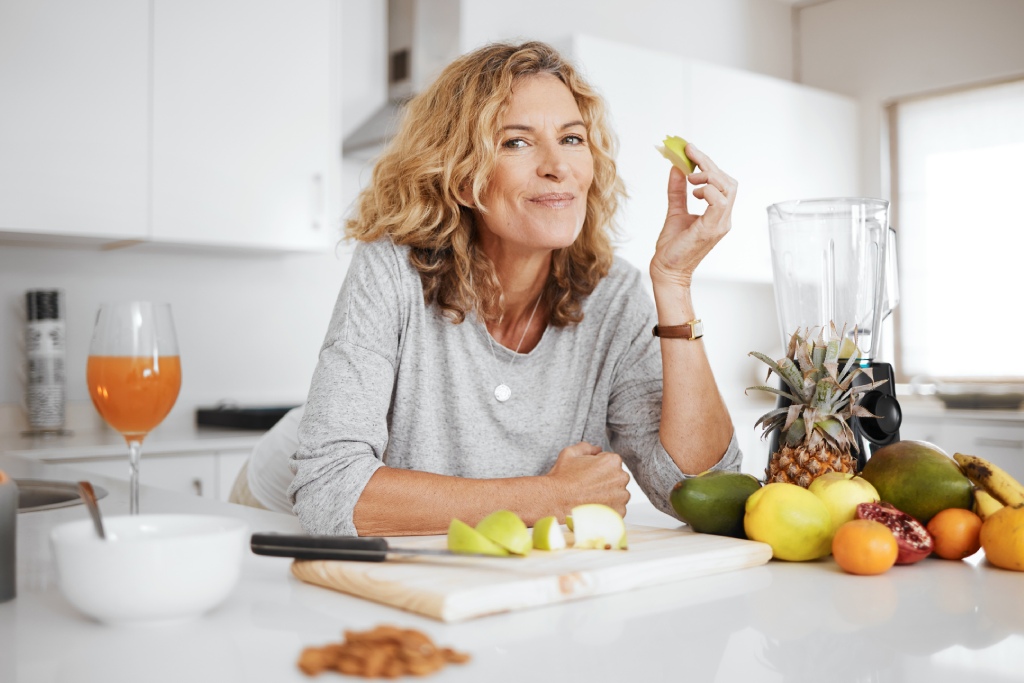 A woman in a kitchen smiles while holding a green apple slice, with fruits and a blender on the counter beside her.