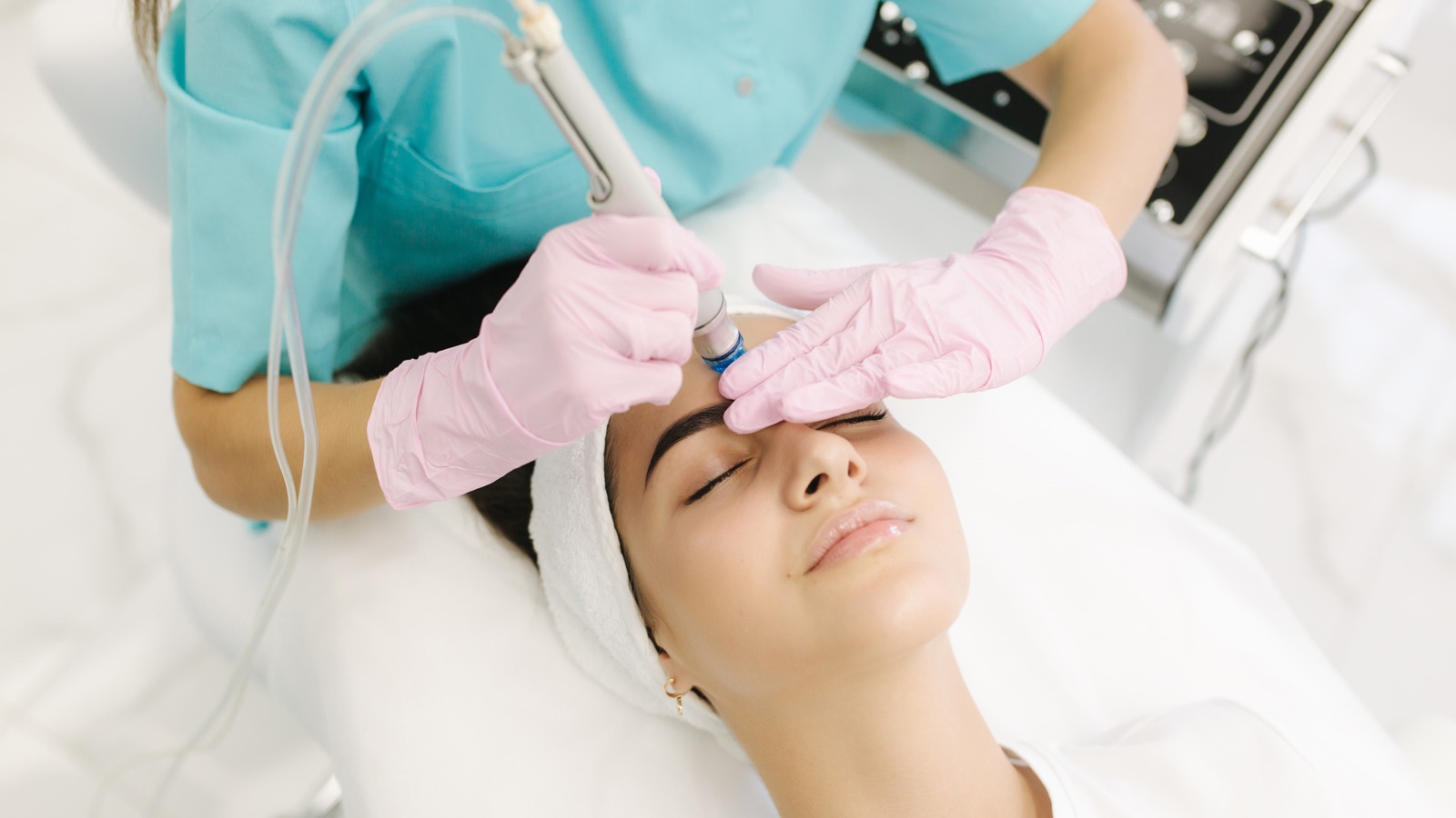 Aesthetician wearing gloves performs a facial treatment using a machine on a relaxed woman lying down with a headband.