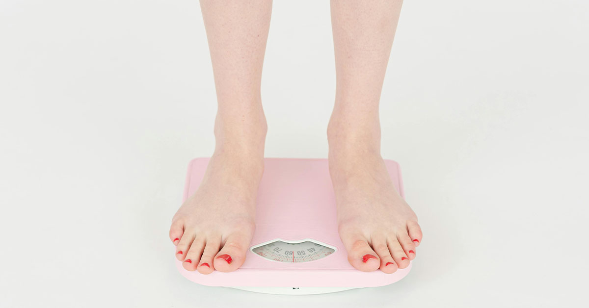 A person with red toenail polish standing on a pink bathroom scale in a medical weight loss clinic against a white background.