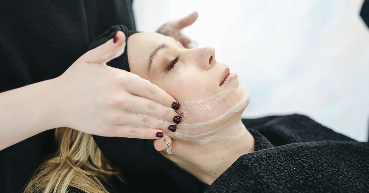 Medspa worker applying skincare facial mask to a relaxed female client lying with eyes closed in a spa setting.