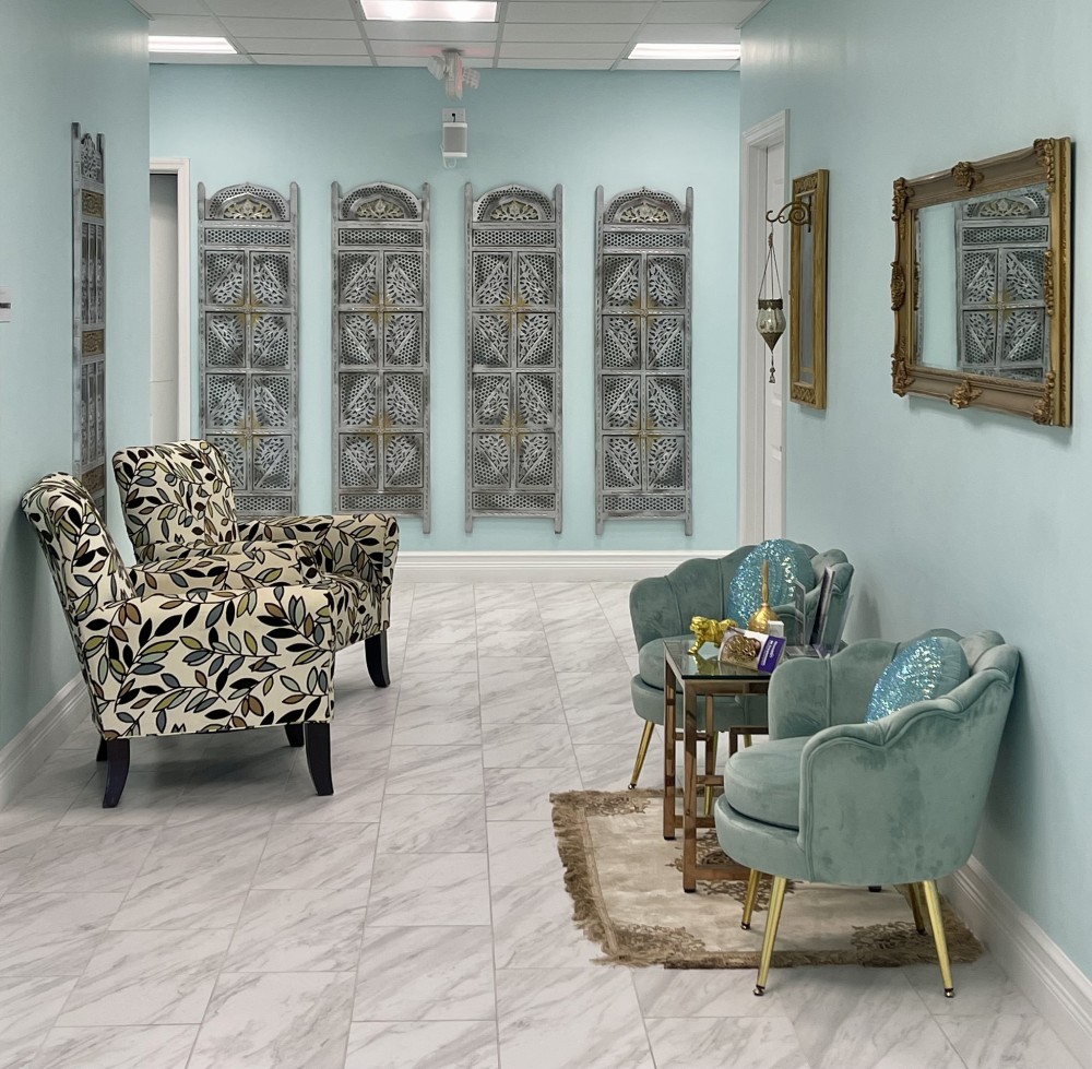 A waiting room with two floral armchairs, three green velvet chairs, a small table with decor items, decorative wall panels, and a gold-framed mirror on light blue walls, and white tiled flooring.